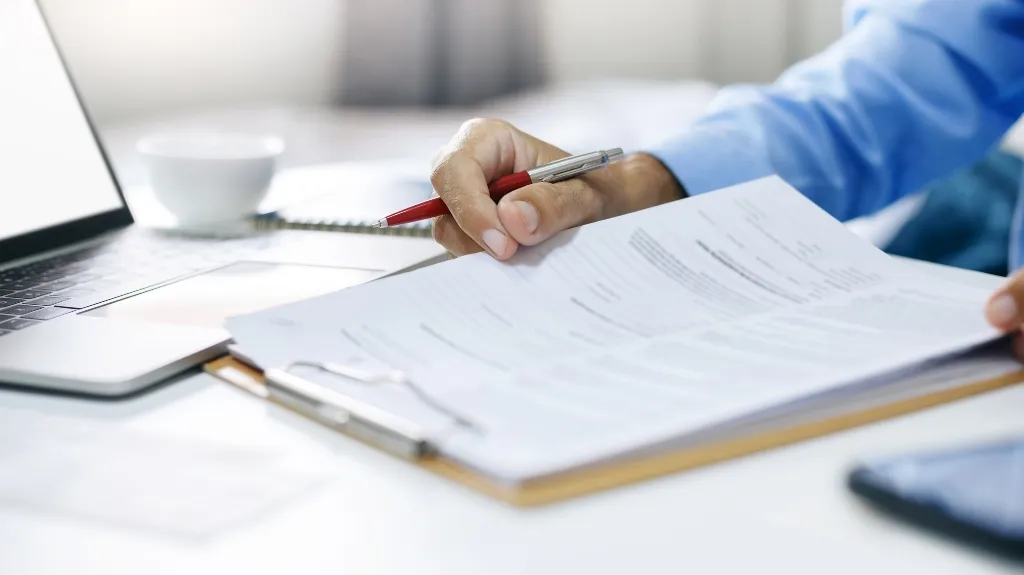 Person in blue shirt holding red pen reviewing documents on clipboard near laptop.