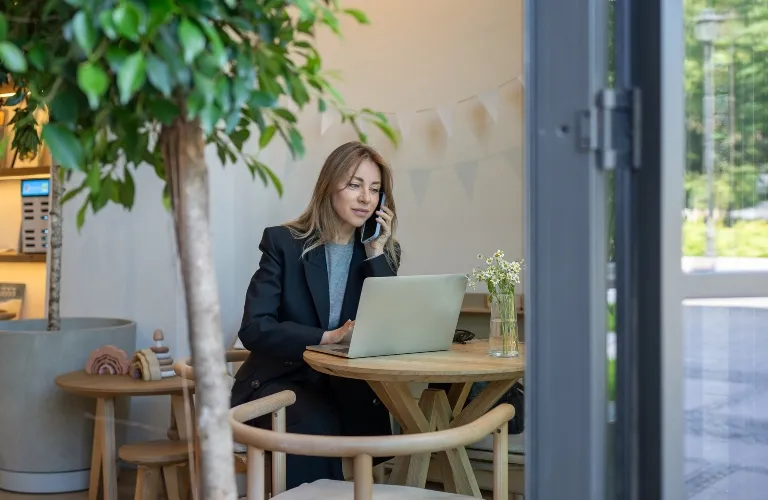 Professional woman in black blazer using laptop and talking on phone at wooden table indoors.