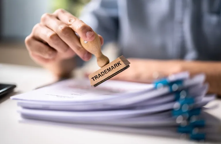 Hand holding a wooden "TRADEMARK" stamp above a stack of white documents with blue clips.