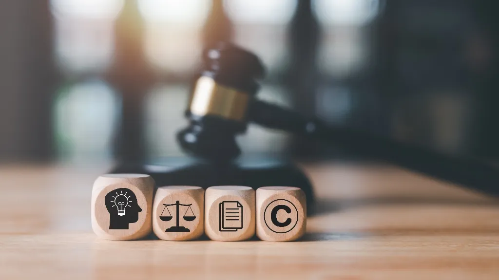 Wooden blocks with intellectual property icons in front of a blurred judge's gavel on wooden surface.